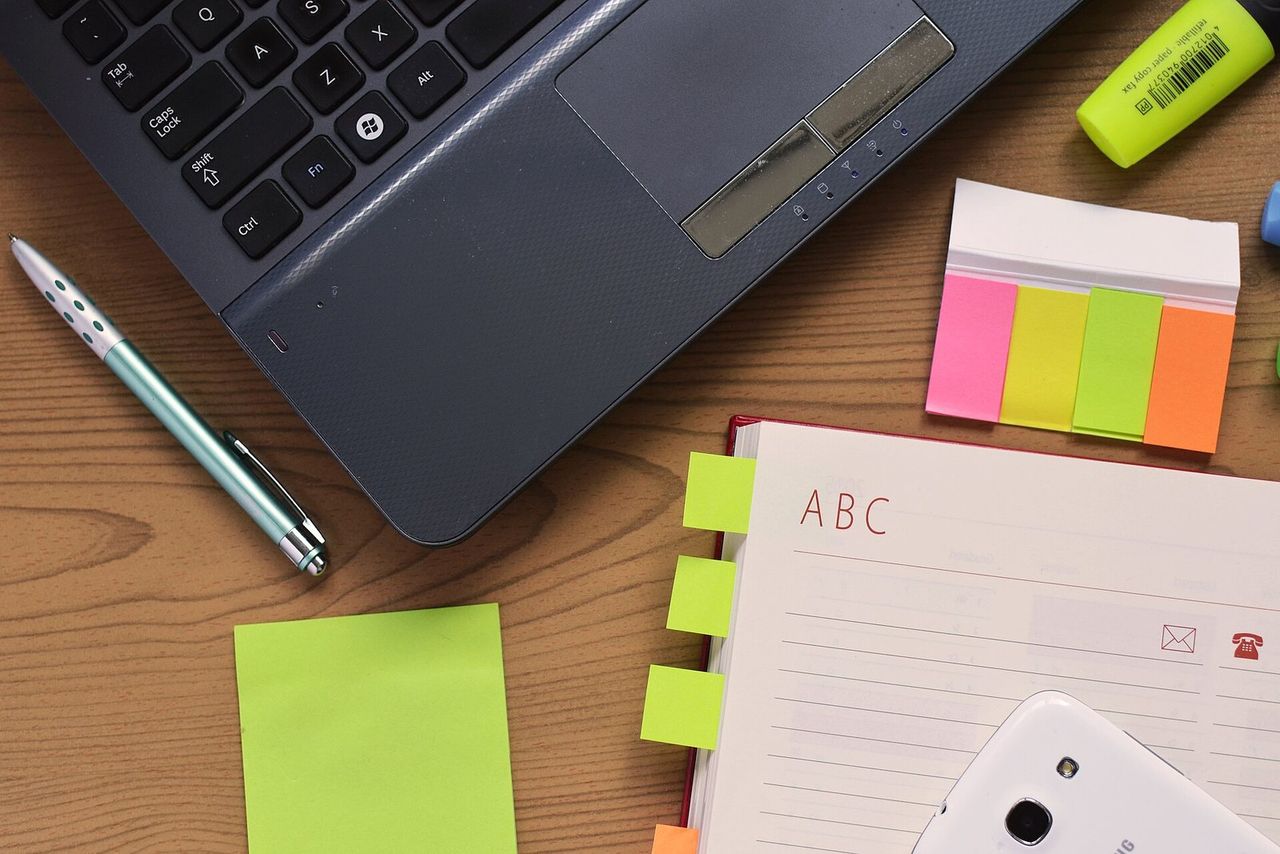 A laptop, lined notebook, pen, and colored sticky notes on a wooden desk